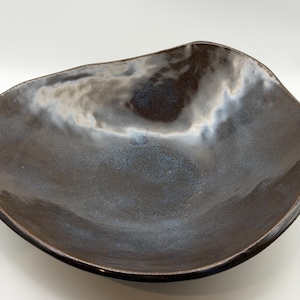 May include: A shallow, hand-formed ceramic bowl with an irregular, organic rim. The surface features a dark, mottled brown glaze with subtle, shimmering blue and silver highlights that create a reflective, metallic-like appearance across the interior.