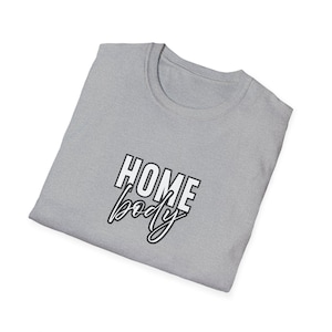 May include: A folded light gray t-shirt with the words "HOME body" printed in black and white. The word "HOME" is in a bold, blocky font, while "body" is in a cursive script. The shirt has a crew neck.