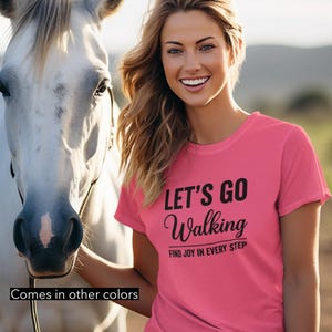 May include: A woman wearing a pink t-shirt with the text "LET'S GO Walking" and "FIND JOY IN EVERY STEP". She is standing next to a white horse. The shirt is paired with blue jeans.