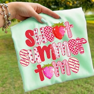 Puede incluir: Una camiseta verde claro con las palabras "Sweet Summer Home" en una fuente lúdica. Las letras son rojas, rosas y blancas, con patrones de fresas y cuadros. También se presentan dos ilustraciones de fresas.