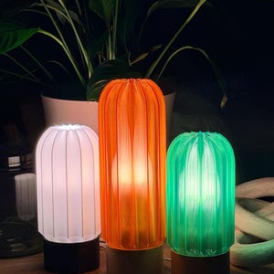 May include: Three decorative table lamps in white, orange, and green. Each lamp has a ribbed, cylindrical shade and a dark brown base. The lamps are illuminated, casting a warm glow. A plant is in the background.