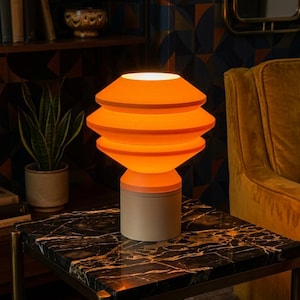 Puede incluir: Una lámpara de mesa naranja escalonada con base beige sobre una mesa auxiliar con tapa de mármol negro y patas doradas. La lámpara está encendida, emitiendo un brillo cálido. El fondo incluye una estantería, una planta y un sillón color mostaza. La lámpara mide unos 30 cm de alto.