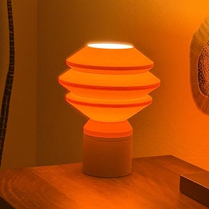 May include: An orange table lamp with a stacked, tiered shade design. The lamp has a cylindrical base and emits a warm, inviting glow. The lamp is sitting on a wooden surface.