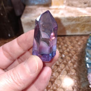 May include: A faceted, purple crystal point with a gradient of colors from deep violet to a lighter, translucent hue. The crystal is held in a hand, showcasing its polished facets and natural beauty. The crystal's shape is a tapered point.