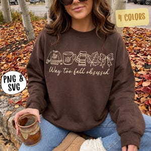 May include: A dark brown crewneck sweatshirt featuring a cream-colored graphic print of a sweater, a mug, a scarf, a leaf, and a bow, with the text "Way too fall obsessed" written in a cursive script below the icons.