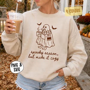 May include: A beige crewneck sweatshirt featuring a brown graphic of a duck wearing a ghost sheet and a plaid scarf while holding a mug. Two small bats are above the duck. The text reads, spooky season, but make it cozy. A yellow tag says 4 COLORS.