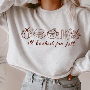 May include: A light grey crewneck sweatshirt featuring a brown line art graphic of a pumpkin, a stack of books, a mug with a pumpkin design, a row of books, and a maple leaf. The text "all booked for fall" is printed in brown cursive script.