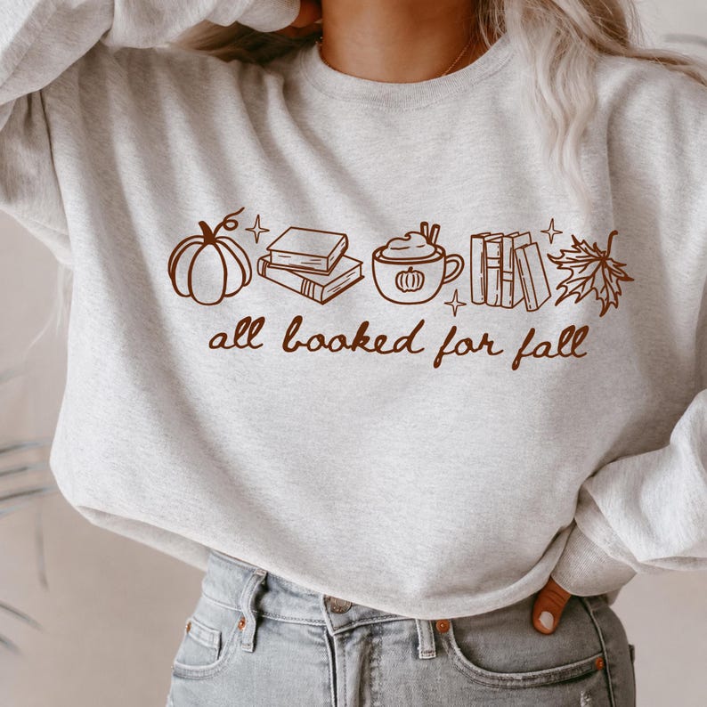 May include: A light grey crewneck sweatshirt featuring a brown line art graphic of a pumpkin, a stack of books, a mug with a pumpkin design, a row of books, and a maple leaf. The text "all booked for fall" is printed in brown cursive script.