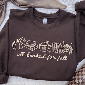 May include: A dark brown crewneck sweatshirt featuring a light-coloured graphic print of a pumpkin, a stack of books, a mug with a pumpkin design, a row of books, and a maple leaf. The text "all booked for fall" is printed below the graphics.