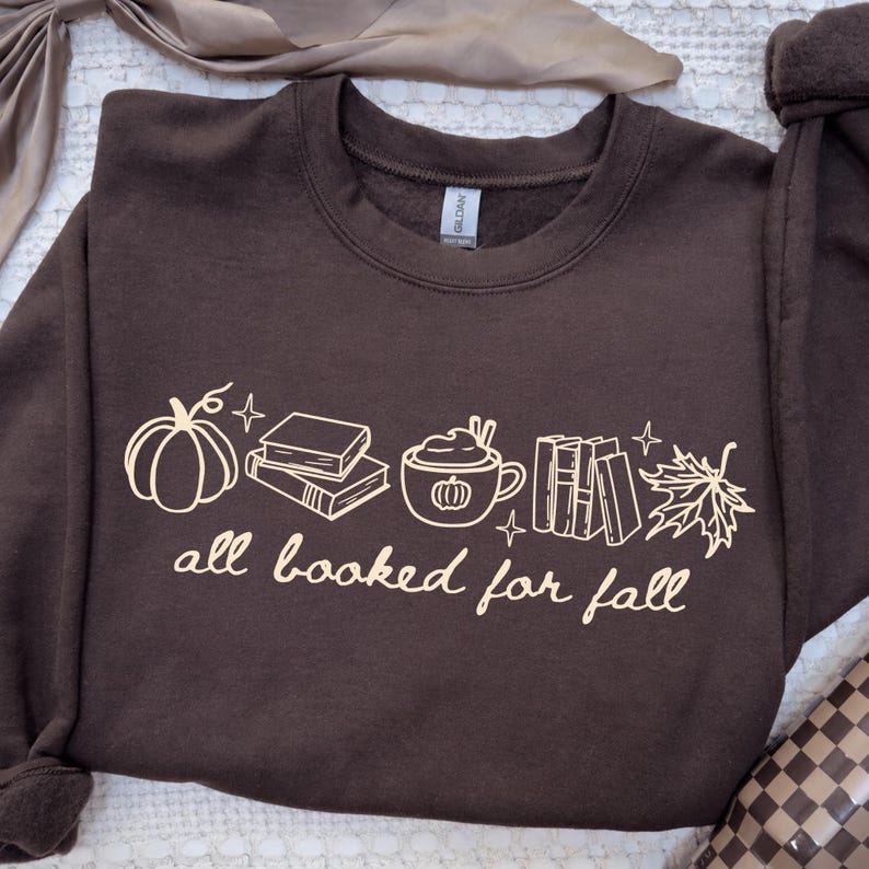 May include: A dark brown crewneck sweatshirt featuring a light-coloured graphic print of a pumpkin, a stack of books, a mug with a pumpkin design, a row of books, and a maple leaf. The text "all booked for fall" is printed below the graphics.