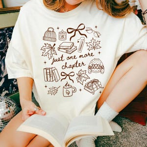 以下が含まれることがあります： クリーム色のTシャツ。ニット帽、キャンドル、リボン、眼鏡、カエデの葉、本、しおり、セーター、マグカップの茶色の線画イラストが描かれています。中央には「just one more chapter」という文字が茶色の筆記体でプリントされています。