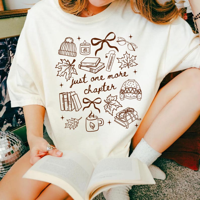 以下が含まれることがあります： クリーム色のTシャツ。ニット帽、キャンドル、リボン、眼鏡、カエデの葉、本、しおり、セーター、マグカップの茶色の線画イラストが描かれています。中央には「just one more chapter」という文字が茶色の筆記体でプリントされています。