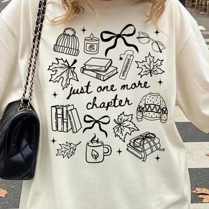 以下が含まれることがあります： 「just one more chapter」という文字と、本、カエデの葉、ニット帽、キャンドル、リボン、眼鏡、セーター、マグカップのイラストが描かれた黒い線画グラフィックがプリントされたクリーム色のTシャツ。