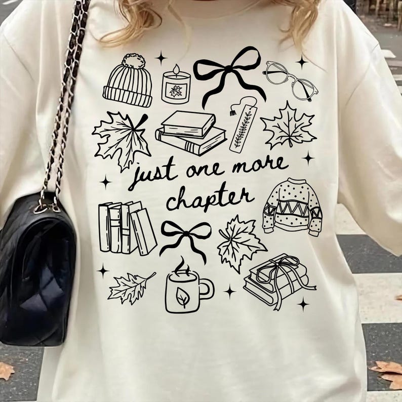 以下が含まれることがあります： 「just one more chapter」という文字と、本、カエデの葉、ニット帽、キャンドル、リボン、眼鏡、セーター、マグカップのイラストが描かれた黒い線画グラフィックがプリントされたクリーム色のTシャツ。