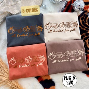Könnte beinhalten: Vier gefaltete Sweatshirts in Dunkelgrau, Creme, Rostorange und Braun. Jedes zeigt eine Strichgrafik mit einem Kürbis, Büchern, einer Tasse und einem Ahornblatt sowie dem Text "all booked for fall" in einer geschwungenen Schreibschrift.