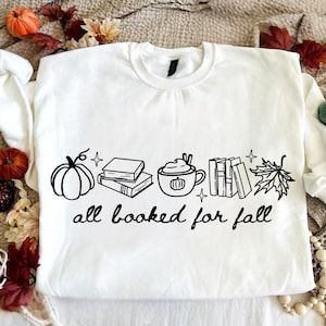 May include: A white long-sleeve sweatshirt featuring a black graphic print of a pumpkin, a stack of books, a mug with a pumpkin design, a row of books, and a maple leaf. The text "all booked for fall" is printed in black below the graphic.