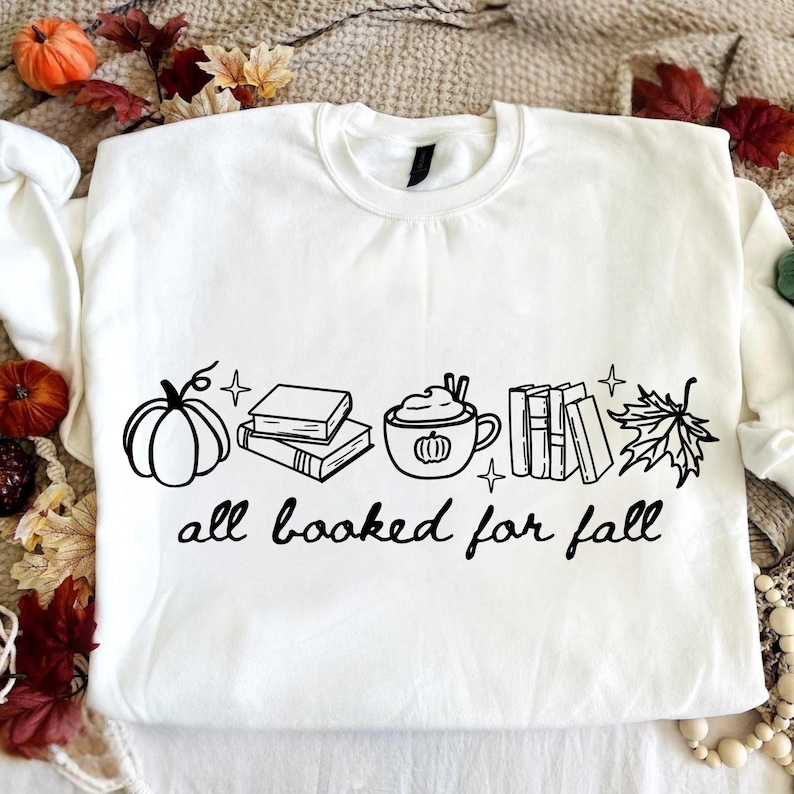 May include: A white long-sleeve sweatshirt featuring a black graphic print of a pumpkin, a stack of books, a mug with a pumpkin design, a row of books, and a maple leaf. The text "all booked for fall" is printed in black below the graphic.