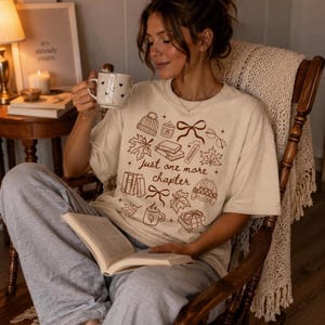 以下が含まれることがあります： クリーム色の半袖Tシャツ。ニット帽、キャンドル、リボン、メガネ、カエデの葉、本、セーターの茶色の線画イラストが描かれています。中央には「just one more chapter」という文字が茶色の筆記体でプリントされています。
