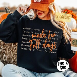 May include: A black long-sleeve sweatshirt featuring white text listing autumn-themed words including Autumn, Leaves, Candles, Cozy, Crisp, Golden, Books, Sweaters, Weather, Latte, Maple, Reading, Pumpkin, Cabin, Pages, Blanket, Fireplace, and November.