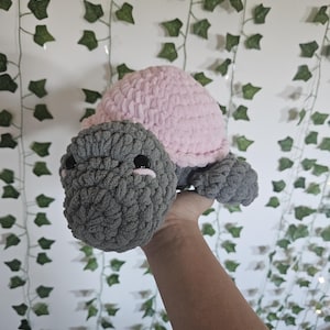 May include: A handmade crochet turtle in shades of pink and gray. The turtle has a pink shell, a gray head, and gray flippers. The eyes are black. The turtle is held up against a white wall with green ivy.