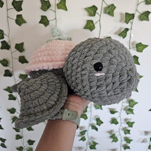 May include: A handmade crochet turtle plush toy featuring a dark gray head and flippers with a soft pink shell. The turtle has two small black plastic eyes and a tiny pink stitched mouth detail, crafted from thick, textured yarn for a soft appearance.