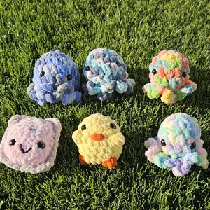 May include: Six handmade crochet plush toys in a variety of colors, including blue, yellow, and pastel shades. The collection features octopus and chick designs, all resting on a bed of green grass.