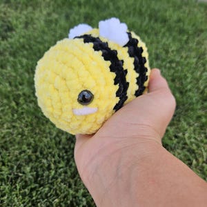 May include: A small, round, crocheted bee plush toy made of soft yellow and black yarn. The bee features two small white yarn wings on its back, a single black plastic safety eye, and a small horizontal white stitched line for a mouth.
