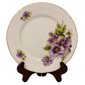May include: White porcelain plate with a scalloped gold rim, adorned with purple and white floral designs and green leaves. The plate is displayed on a dark brown wooden stand.