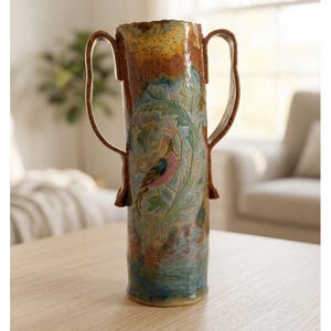 May include: A tall, cylindrical ceramic vase with two large, curved handles. The surface features a textured, multi-colored glaze in shades of brown, blue, and green, with an embossed design of a bird perched among leafy branches on the front.