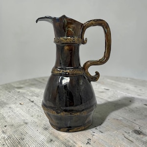 May include: A ceramic pitcher with a glossy dark brown and black glaze. The vessel features a wide, rounded base, a narrow neck, and a curved handle with decorative textured bands around the neck and body, finished with a pointed pouring spout.