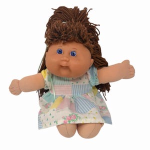 May include: A vintage doll with brown yarn hair and blue eyes. The doll wears a patchwork dress with pastel colors and floral patterns. The doll's arms are outstretched, and it has a soft, plush body.