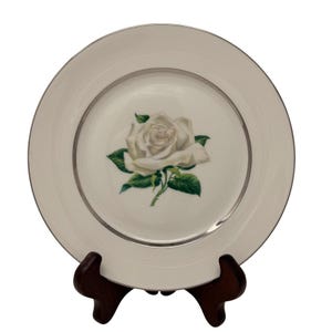 May include: A white porcelain plate with a silver rim and a central image of a white rose with green leaves. The plate is displayed on a dark brown wooden stand.