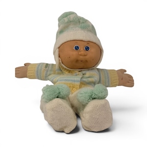 May include: A vintage Cabbage Patch Kid doll, seated with arms outstretched. The doll wears a knit outfit in cream, yellow, and blue stripes, with a matching hat. The doll's feet have green pom-poms, and it has blue eyes and a painted face.