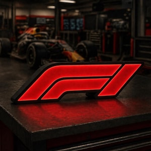 Puede incluir: Un logotipo de F1 rojo y negro iluminado, probablemente un artículo decorativo. El logotipo se ilumina con un brillo rojo brillante, sobre un fondo borroso de un coche de carreras y un taller.