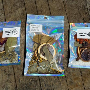 May include: Three iridescent holographic pouches containing dried botanical ingredients for simmer pots. The labels read Summer Simmer Pot, Breathe Babe Anti-Funk Simmer Pot, and Velvet Sun Simmer. Each pouch holds dried citrus slices, leaves, and herbs.