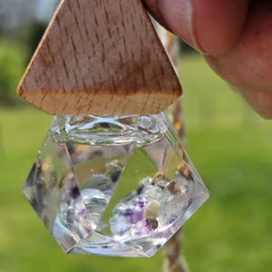 May include: A clear, geometric pendant with a wooden triangle top. The pendant contains small, colorful stones and is suspended by a natural cord. The background is blurred green.