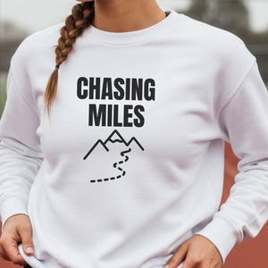 May include: White sweatshirt featuring the words "CHASING MILES" in black, accompanied by a mountain and dotted path graphic. The sweatshirt has long sleeves and a crew neck. The person is wearing black shorts.