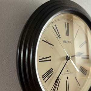以下が含まれることがあります： 黒いフレームとクリーム色の文字盤の、丸いセイコーの壁掛け時計。 時計には黒いローマ数字と針があり、「SEIKO」と「THE NATIONAL TRUST」の文字が見えます。 時計は明るい灰色の壁に取り付けられています。