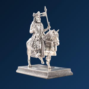 May include: A silver-colored metallic figurine depicting a royal figure wearing a crown and traditional attire, mounted on a horse. The figure holds a tall spear with a flag. The entire sculpture is set upon a rectangular base with a textured pattern.