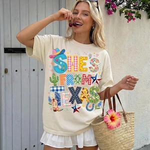May include: Cream-colored t-shirt with the text "SHE'S FROM TEXAS" in colorful, patchwork-style letters and designs. The design includes a cowboy hat, cactus, stars, and a horseshoe. A straw tote bag with a pink flower.