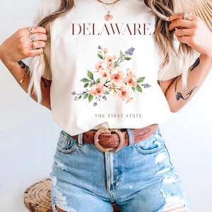 May include: Cream-colored t-shirt featuring the word "DELAWARE" in burgundy above a floral design with peach and purple flowers. The text "THE FIRST STATE" is below the floral design. The model is wearing blue denim shorts and a brown belt.