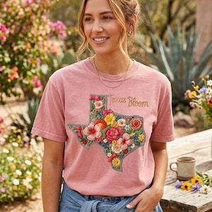 Puede incluir: Camiseta de color rosa polvoriento con un diseño floral del contorno de Texas. El diseño bordado incluye varias flores coloridas. Las palabras "Texas Bloom" están escritas encima del diseño en letras doradas. La camiseta se combina con una chaqueta vaquera.