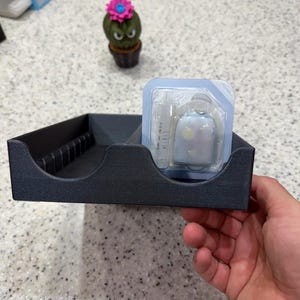 Può includere: Un vassoio portaoggetti in plastica grigio scuro con una serie di fessure verticali sul lato sinistro e uno scomparto rettangolare a destra contenente un dispositivo medico in blister trasparente con una siringa e un piccolo contenitore bianco.