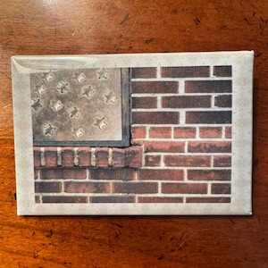 May include: Rectangular decorative item featuring a patriotic design. The image depicts a section of a brick wall and a metal panel with star cutouts, resembling a flag. The item has a neutral-toned border.