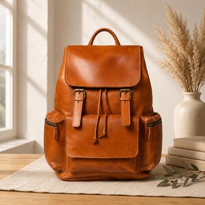 May include: A cognac-coloured leather backpack featuring a top flap with two buckle straps, a drawstring closure, and two side pockets with zip closures. The bag includes a top handle and a large front pocket with a flap closure.