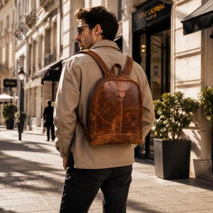 May include: A brown leather backpack with a smooth, distressed finish, featuring a rounded top, a vertical center seam, and adjustable shoulder straps. The bag has a structured shape with a top handle and a zippered main compartment closure.
