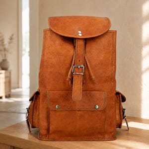 May include: A tan leather backpack featuring a large top flap with a central vertical strap and a silver metal buckle. The bag includes a front pocket with two snap buttons and two side pockets, all constructed from smooth, durable tan leather.