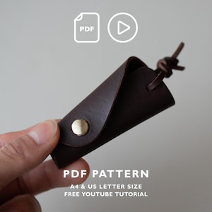 May include: A dark brown leather key holder featuring a gold-tone metal snap button closure and a leather cord tie. The image includes icons for PDF and video, with text reading PDF PATTERN, A4 & US LETTER SIZE, FREE YOUTUBE TUTORIAL.