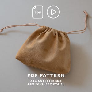 May include: A tan suede drawstring pouch with thin matching cords. Above the pouch are white icons for a PDF file and a play button. Text below reads: PDF PATTERN, A4 & US LETTER SIZE, FREE YOUTUBE TUTORIAL. The pouch has a gathered top closure.