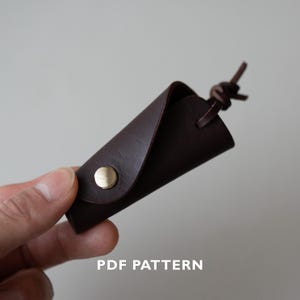 May include: A dark brown leather key holder with a brass snap closure. The key holder is folded into a triangular shape and has a leather loop with a knot at the top. The text "PDF PATTERN" is at the bottom.